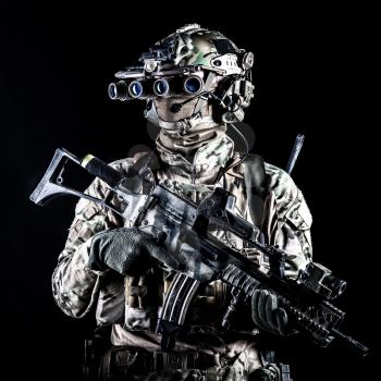 Marine rider in camouflage uniform and face mask, patrolling in darkness with quad-tube night vision goggles on battle helmet, holding modern assault rifle, low key studio portrait on black background