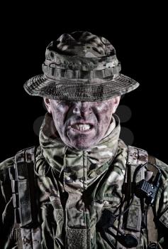 Commando fighter, elite troops soldier in camo uniform, bending head, hiding eyes behind bonnie hat, clenching teeth with anger and aggression, low key, studio portrait isolated on black background