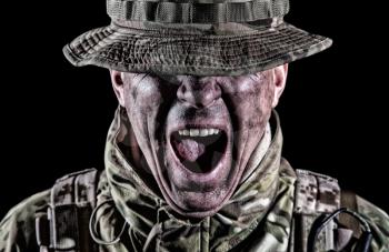 Commando fighter, elite troops soldier in camo uniform, bending head, hiding eyes behind bonnie hat, clenching teeth with anger and aggression, low key, studio portrait isolated on black background