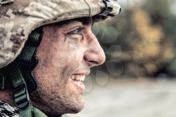 Shoulder portrait of happy smiling young soldier in battle helmet with scratches on camouflage, equipped U.S. army contractor, Marine Corps infantryman with dirty face looking at camera with friendly smile
