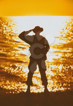 Silhouette of saluting commando soldier, army infantryman standing on shore during sunset or sunrise. Military solemn ceremony, respectable salute for honoring fallen heroes and comrades on seacoast