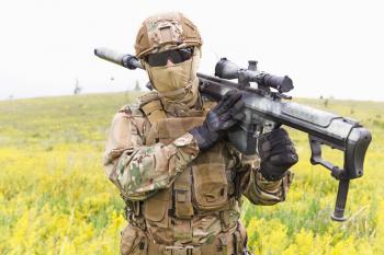 Special forces soldier walks across the yellow field with sniper rifle close up
