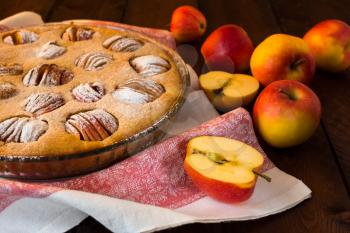 Apple pie in a baking dish, linen napkin and apples on dark background, selective focus