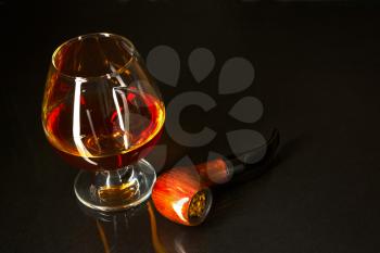 Whiskey glass and smoking pipe on black background. Cognac glass. Brandy glassful. Cognac france. Scotch drink. Smoking pipe.