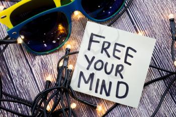 Free your Mind encouragement concept on note paper. Some lights are shown in discrete order. Two sunglasses of discrete colors with wooden background. Positive Inspiration concept words.
