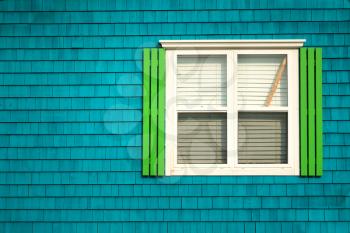 Traditional window with neon green shutters on a dark turquoise wooden tiles wall in Iles de la Madeleine in Quebec, Canada