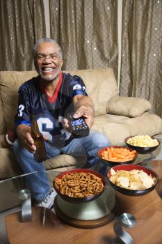 Portrait of a Middle-aged African-American man wearing a football jersey surrounded by snacks pointing a remote at viewer.