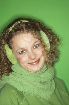 Portrait of smiling young adult Caucasian woman on green background wearing earmuffs, winter coat and scarf.