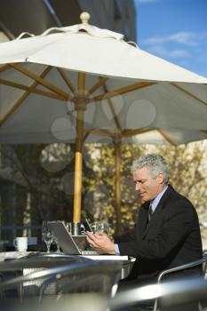 Royalty Free Photo of a Businessman Sitting at a Patio Table Outside With Laptop and Dialing on His Cellphone