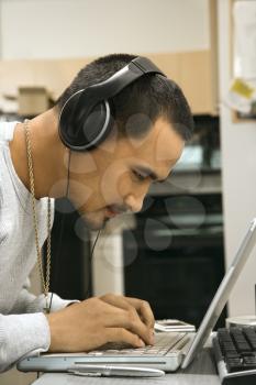 Close-up side view of Asian young adult man leaning over laptop typing on keyboard wearing headphones.