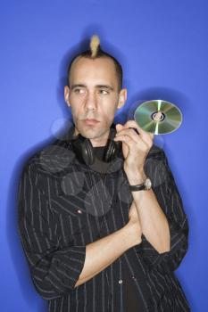 Caucasian man with mohawk with headphones holding cd standing against blue background.