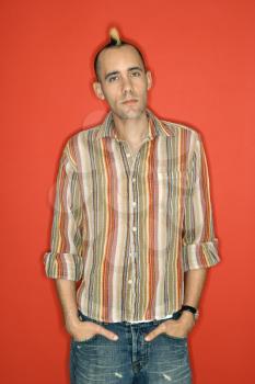 Caucasian man with mohawk standing against red background.