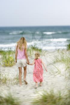 Caucasian pre-teen girl holding hands with younger Caucasian girl walking toward beach.