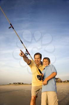 Caucasian mid-adult man shore fishing on beach with pre-teen boy and pointing.