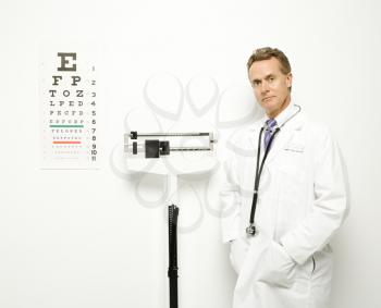 Mid-adult Caucasian male doctor standing next to scale with eye chart on wall.