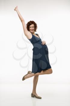Royalty Free Photo of an Attractive Pregnant Woman Standing With One Leg Up, Hand on Belly and Arm in Air Smiling