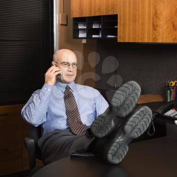 Royalty Free Photo of a Middle-Aged Businessman in an Office Sitting With His Feet on a Desk Talking on a Cellphone
