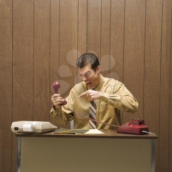 Caucasion mid-adult retro businessman sitting at desk pointing at telephone in anger.