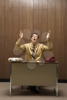 Royalty Free Photo of a Businessman Sitting at a Desk Raising Hands in the Air With a Look of Frustration