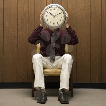 Royalty Free Photo of a Middle-aged Businessman Holding a Clock in Front of His Head While Sitting in Chair Against Wood Paneling