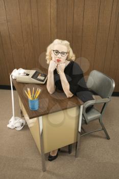 Mid-adult Caucasian female in vintage outfit sitting at desk with chin on her fists.