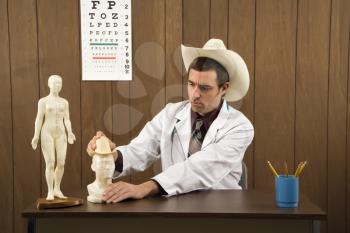 Royalty Free Photo of a Male doctor wearing cowboy hat sitting at desk playing with figurine