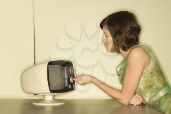 Royalty Free Photo of a Side View of a Woman Wearing a Green Vintage Dress Sitting at s 50's Retro Dinette Set Turning an Old Television Knob