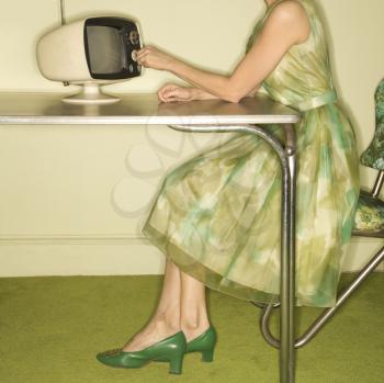 Royalty Free Photo of a Side View of a Woman Wearing a Green Vintage Dress Sitting at s 50's Retro Dinette Set Turning an Old Television Knob