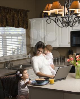 Royalty Free Photo of a Mother Holding Her Baby and Typing on a Laptop While Daughter Eats Breakfast