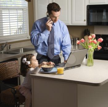 Royalty Free Photo of a Father in a Suit Talking on a Cellphone and Using a Laptop Computer With Daughter Eating Breakfast in the Kitchen