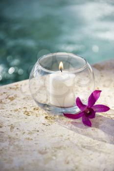 Lit candle in glass bowl with purple orchid next to pool.