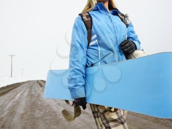 Royalty Free Photo of a Woman Walking Alone Down a Muddy Dirt Road Holding a Snowboard and Boots