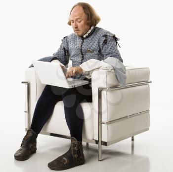 William Shakespeare in period clothing sitting on modern chair using laptop.