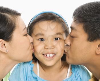 Asian mother and father kissing opposite cheeks of smiling daughter in front of white background.
