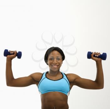 African American young adult woman raising dumbbells over head and smiling at viewer.