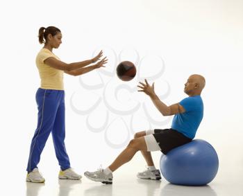 Mid adult multiethnic woman throwing ball to multiethnic mid adult man balancing on blue excersise ball.
