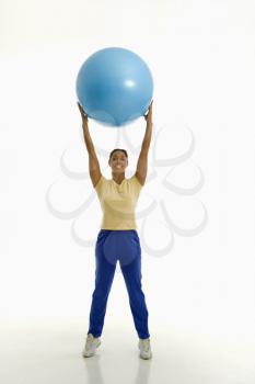 Mid adult multiethnic woman standing and holding blue exercise ball over her head looking at viewer and smiling.