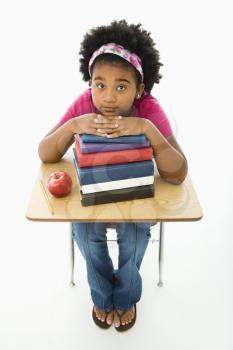 African American girl sitting at school desk with large stack of books looking up at viewer.