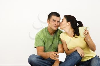 Couple sitting on floor drinking coffee with woman leaning over kissing man on cheek.