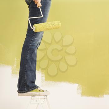 Royalty Free Photo of a Woman's Legs Standing on a Stepladder Holding a Paint Roller Beside a Painted Wall