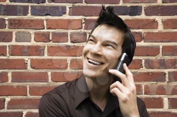 Smiling young Asian man next to brick wall talking on cell phone.