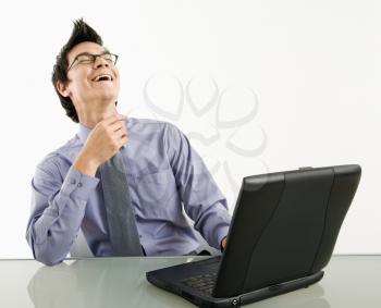 Asian businessman sitting at desk working on laptop computer laughing.