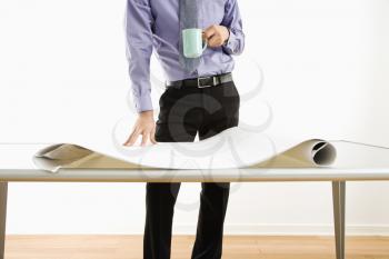 Business man standing with coffee cup next to table with blueprints.