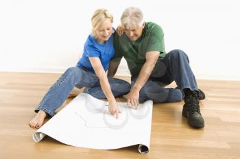 Middle-aged couple sitting on floor looking at and discussing architectural blueprints together.