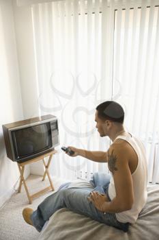 Young Asian man sitting on the edge of a bed and turning on the television. Vertical shot.