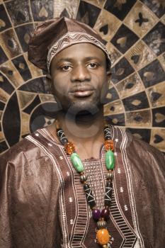 Portrait of an African American man wearing traditional African clothing, in front of a patterned wall and looking at the camera. Vertical format.