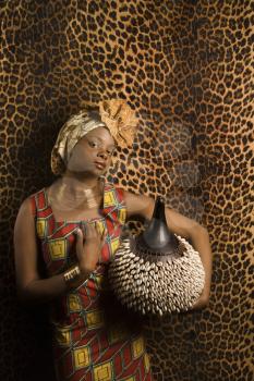Portrait of an African American woman wearing traditional African clothing and holding a shekere in front of a patterned wall. Vertical format.