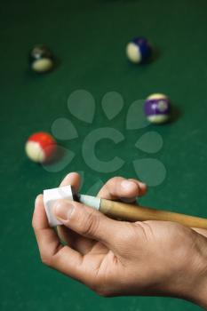 Hand chalking a pool cue with a billiards table in background. Vertical shot.