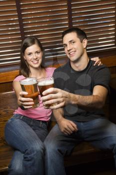 Young man and woman sitting together toasting their beers at pub looking at viewer. Vertical shot.