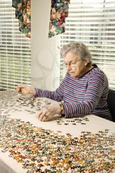 Elderly woman sitting at table sorting through a jigsaw puzzle pieces. Vertical shot.